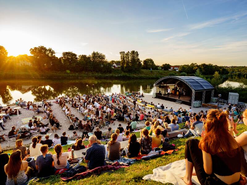 Neckarstrand in Remseck mit untergehender Sonne, dem Neckar im Hintergrund und zahlreichen Menschen, die auf eine Freilichtbühne schauen