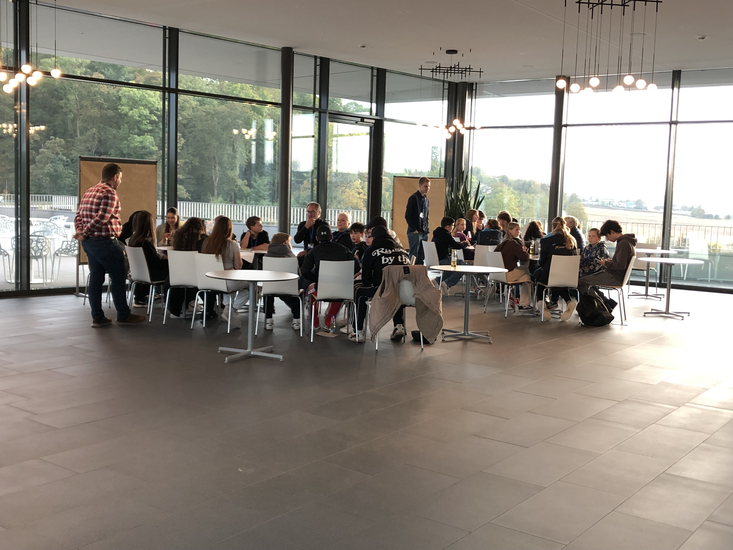 Zwei Gruppen von Menschen, die sich um zwei Tische versammelt haben und gemeinsam an etwas arbeiten, während eine Person daneben steht und zuschaut.