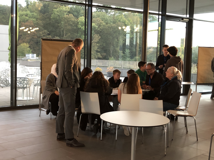 Eine Gruppe von Menschen, die sich um einen Tisch versammelt hat und gemeinsam an etwas arbeitet, während eine Person daneben steht und zuschaut.