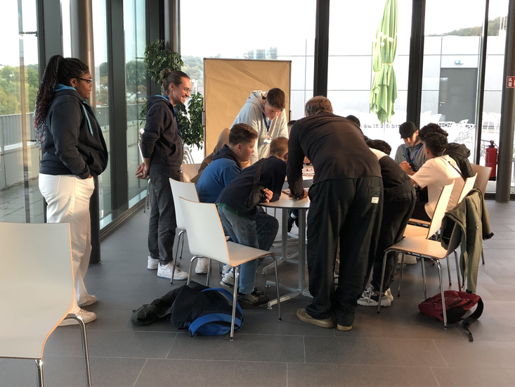 Eine Gruppe von Menschen, die sich um einen Tisch versammelt hat und gemeinsam an etwas arbeitet, während eine Person daneben steht und zuschaut.