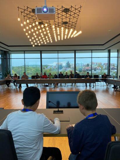 Zwei Kinder an einem Tisch mit Mikrofon in einem Sitzungssaal. Weitere Kinder in den Zuschauerrängen.