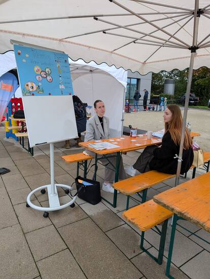 Zwei Personen sitzen unter einem Pavillon an einem Tisch mit Informationsmaterialien, neben einem Plakatständer und einem Schild mit der Aufschrift „Remsos kleine Weltretter“.