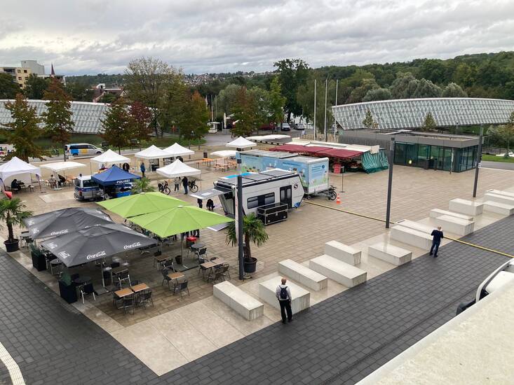 Ein Platz mit mehreren Pavillons und Verkaufsständen, umgeben von Bäumen und einem großen, modernen Gebäude im Hintergrund.