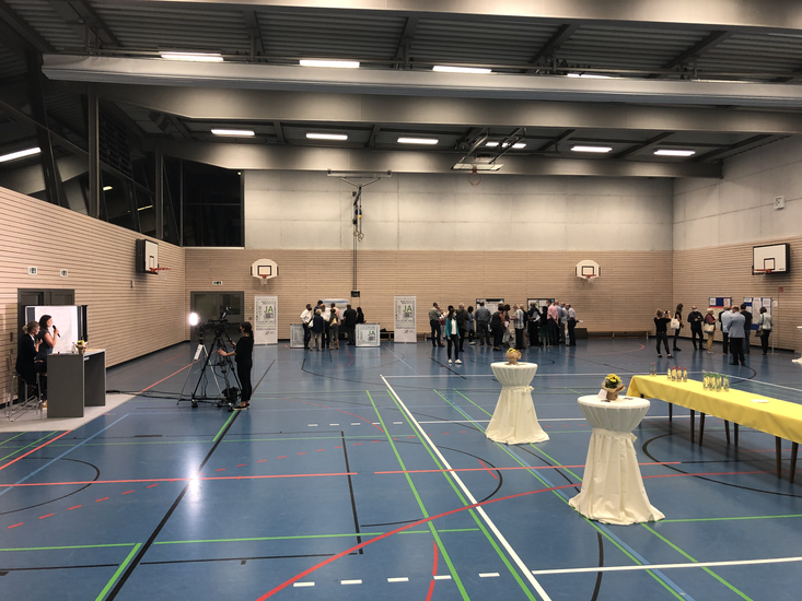 In einer großen Sporthalle mit blauen Bodenlinien und mehreren Basketballkörben findet eine Versammlung von Menschen statt, während im Vordergrund Stehtische und ein gelb gedeckter Tisch mit Getränken zu sehen sind.