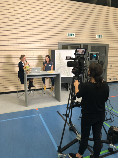 Zwei Frauen sitzen an einem Tisch und sprechen in Mikrofone, während eine Kamerafrau sie filmt.