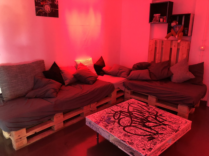 Jugendraum mit Palettenmöblen, rotem gedimmtem Licht und einem kleinen Couchtisch in der Mitte.