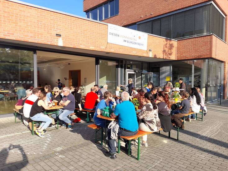 Menschen an Biertischgarnituren vor einem Gebäude.