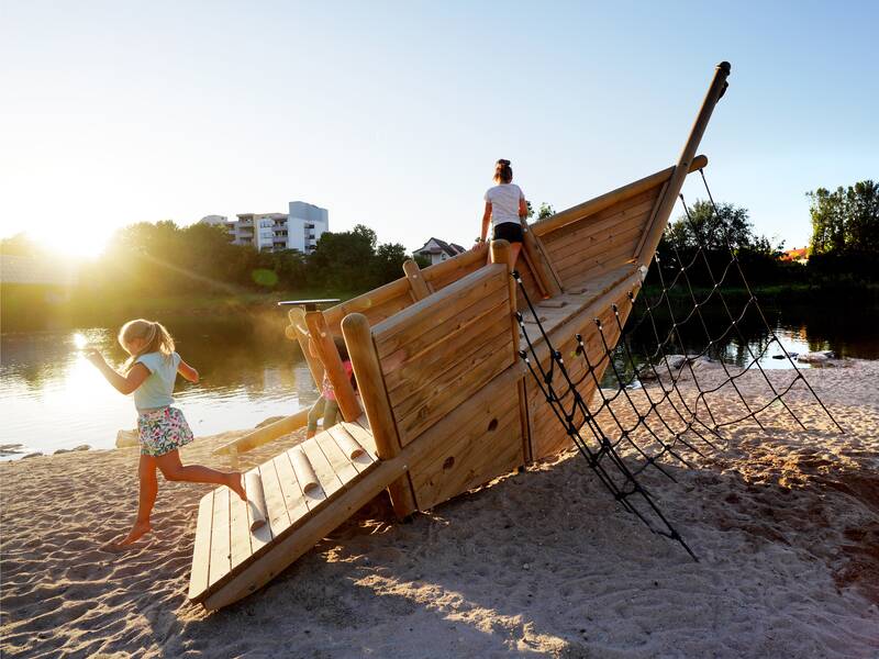 Ein Holzschiff als Spielgerät für Kinder am Neckarstrand bei Sonnenschein. Zwei Kinder spielen.