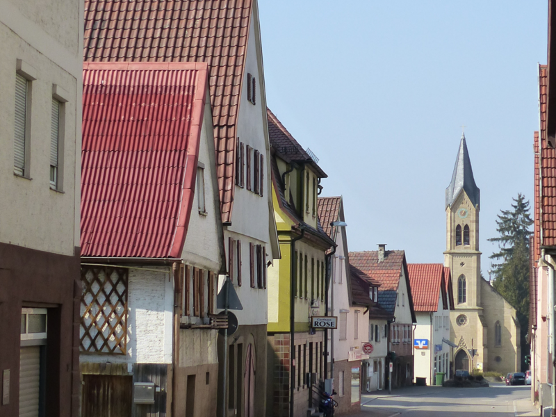 Eine schmale Straße in einem malerischen Dorf mit Fachwerkhäusern und einer Kirche mit Turm im Hintergrund.