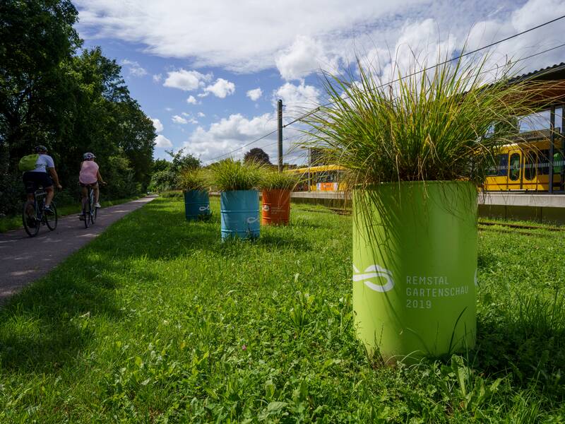 Radweg mit zwei Fahrradfahrern und alte besprühte und bepflanzte Ölfässer mit der Aufschrift: Remstal Gartenschau 2019.