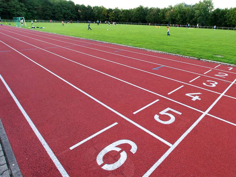 Eine Laufbahn mit sechs Bahnen erstreckt sich entlang eines Sportfeldes, auf dem Menschen Fußball spielen, umgeben von Bäumen.