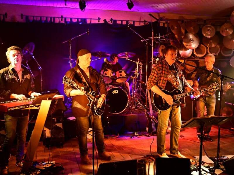 Eine Band spielt auf einer Bühne, beleuchtet von bunten Lichtern, mit einem Keyboarder, zwei Gitarristen, einem Bassisten und einem Schlagzeuger.