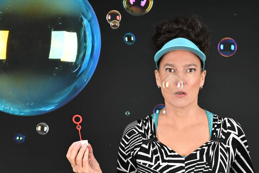 Eine Frau mit einem visierartigen Stirnband pustet vor einem dunklen Hintergrund Seifenblasen, von denen eine direkt vor ihrem Gesicht schwebt.