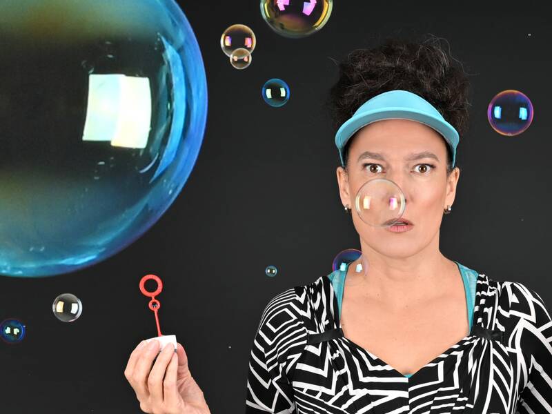 Eine Frau mit einem visierartigen Stirnband pustet vor einem dunklen Hintergrund Seifenblasen, von denen eine direkt vor ihrem Gesicht schwebt.