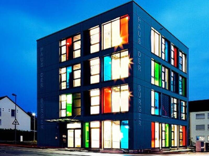 Ein modernes, würfelförmiges Gebäude mit mehreren Stockwerken, dessen Fenster in verschiedenen Farben erleuchtet sind, steht in einer städtischen Umgebung bei Dämmerung.