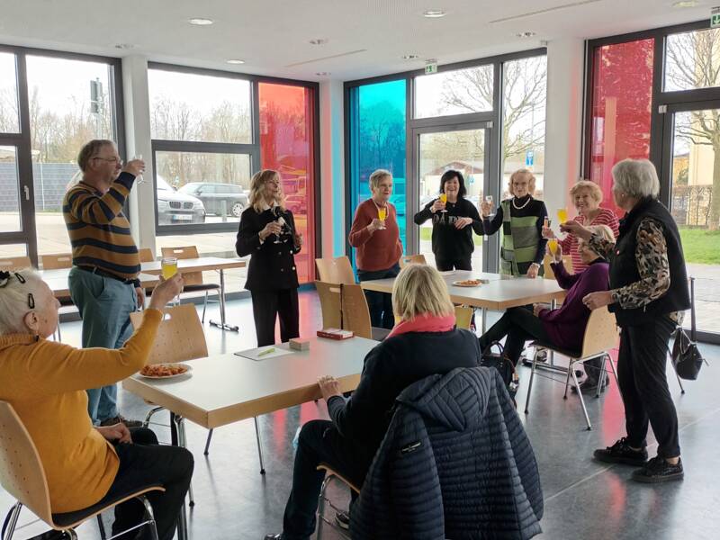 Eine Gruppe von Menschen steht und sitzt an einem Tisch in einem Raum mit bunten Fenstern und stößt vermutlich mit Getränken an.