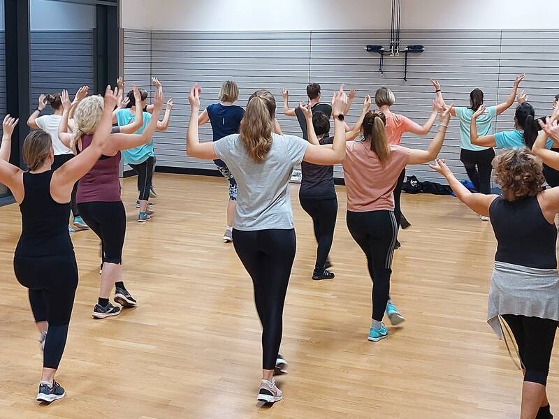 Eine Gruppe von Menschen nimmt in einem Fitnessstudio an einem Tanzkurs teil und hebt synchron die Arme.