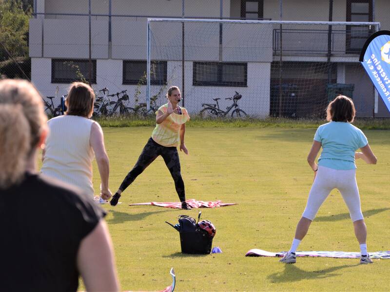Mehrere Personen nehmen an einem Fitnesskurs auf einer Wiese teil, angeleitet von einer Frau in sportlicher Kleidung.