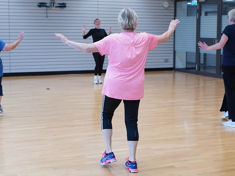 Vier Personen mittleren Alters führen in einem hellen Sportraum synchron eine Fitnessübung aus.