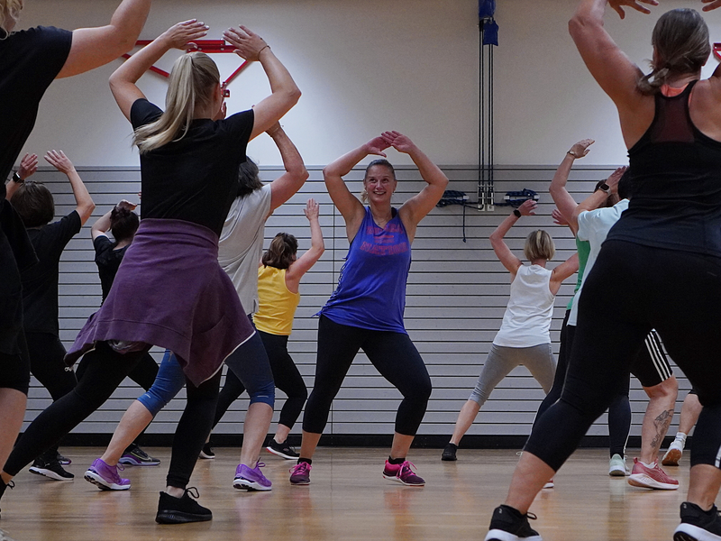Eine Gruppe von Menschen nimmt in einem Fitnessstudio an einem Tanz- oder Aerobic-Kurs teil, wobei alle synchron mit erhobenen Armen üben.