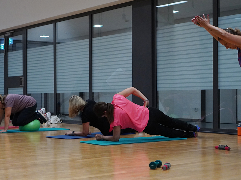 In einem Fitnessraum machen mehrere Personen verschiedene Übungen auf Matten, während eine Trainerin Anweisungen gibt.