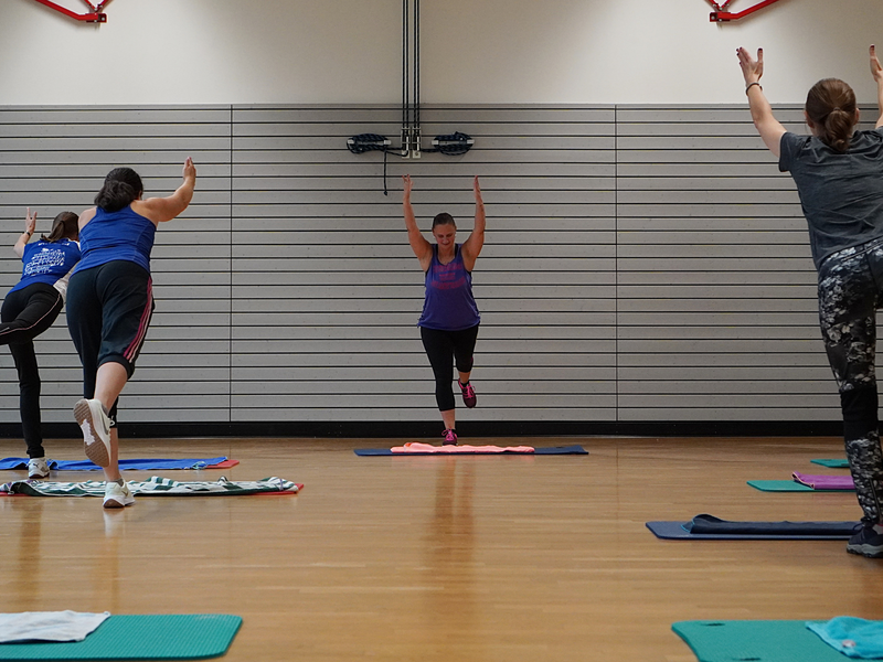 In einem Fitnessstudio machen mehrere Personen gemeinsam Yoga-Übungen auf Matten, wobei sie ein Bein anheben und die Arme in die Höhe strecken.