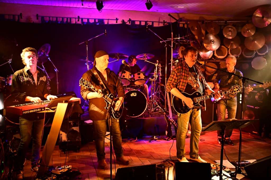Eine Band spielt auf einer Bühne, beleuchtet von bunten Lichtern, mit einem Keyboarder, zwei Gitarristen, einem Bassisten und einem Schlagzeuger.